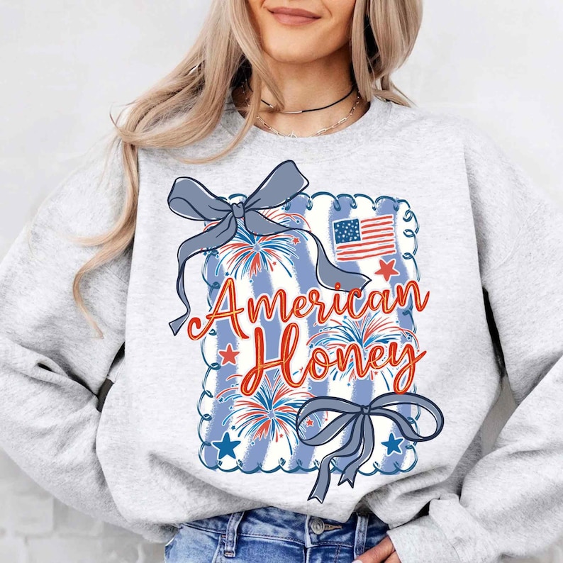May include: A light grey sweatshirt with a patriotic design. The design features the text "American Honey" in red script, a blue and white striped background, fireworks, stars, and a small American flag. Two grey bows are also included in the design.