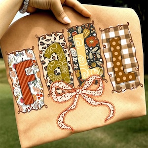 May include: A tan-colored tote bag with the word "FALL" in decorative letters. The letters are filled with various patterns, including floral, leopard print, and plaid. A dotted bow is at the bottom.