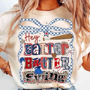 Può includere: T-shirt color crema con grafica a tema baseball. Il design presenta le parole "Hey Batter Batter Swing" in un mix di font e motivi, tra cui stelle, palline da baseball e una mazza da baseball. Un fiocco a scacchi blu e bianco incornicia il design.