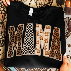 May include: Black sweatshirt with the word "MAMA" in large, decorative letters. The letters have different patterns: leopard print, black and white checkerboard, and a cream color with orange pumpkin and bow designs. The sweatshirt is held by hands.