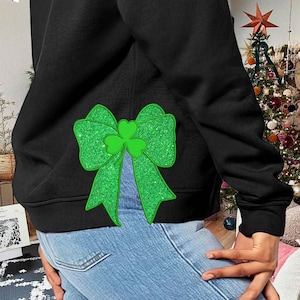 May include: Black hoodie with a green glitter bow and shamrock design on the back. The hoodie is worn over blue jeans.