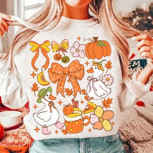 May include: White sweatshirt featuring a fall-themed design. The design includes orange bows, pumpkins, flowers, ducks, and autumn leaves. The illustration has a retro, cartoonish style with a warm color palette.