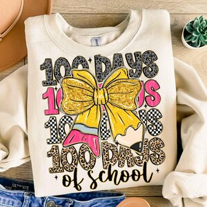 Puede incluir: Sudadera de color crema con un gr&aacute;fico que celebra los 100 d&iacute;as de escuela. El dise&ntilde;o incluye el texto "100 Days of School", un gran lazo dorado con purpurina, un l&aacute;piz y detalles en rosa. La sudadera est&aacute; doblada sobre una superficie de madera.