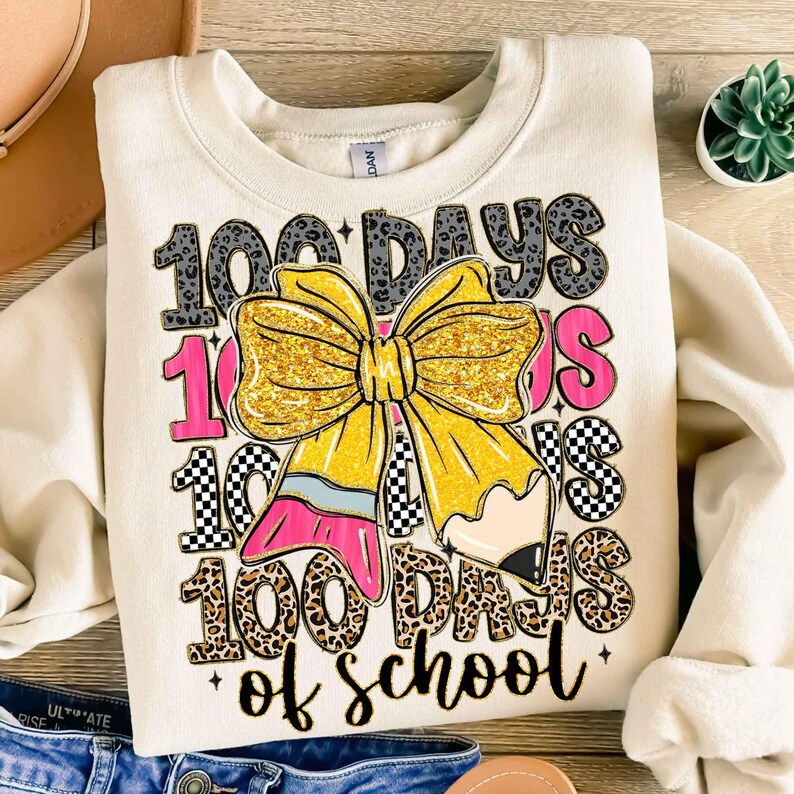 Puede incluir: Sudadera de color crema con un gr&aacute;fico que celebra los 100 d&iacute;as de escuela. El dise&ntilde;o incluye el texto "100 Days of School", un gran lazo dorado con purpurina, un l&aacute;piz y detalles en rosa. La sudadera est&aacute; doblada sobre una superficie de madera.