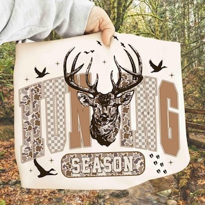 K&ouml;nnte beinhalten: Cremefarbene Grafik mit einer detaillierten Illustration eines Hirschkopfes mit gro&szlig;em Geweih. Das Wort "HUNTING" steht &uuml;ber dem Hirsch, und "SEASON" darunter. Das Design enth&auml;lt Tarn- und Schachbrettmuster mit fliegenden V&ouml;geln.