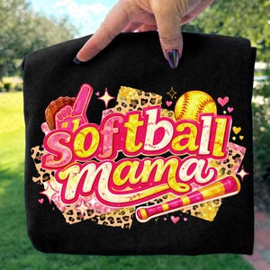 Può includere: T-shirt nero con la scritta "Softball Mama" in un design colorato. Il disegno include una palla da softball, una mazza, un guanto e stelle, con dettagli rosa, gialli e stampa leopardata. La maglietta è tenuta da una mano.