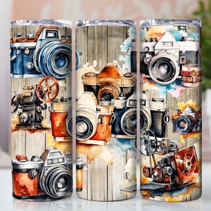 Puede incluir: Tres vasos de acero inoxidable con un diseño de acuarela que presenta cámaras vintage. Los vasos tienen un fondo de veta de madera con salpicaduras de color.