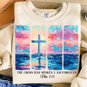 May include: A beige sweatshirt with a colorful painting of a cross over a blue ocean and a pink and blue sky. The text "The cross has spoken, I am forgiven 1 Peter 2:24" is printed below the painting.