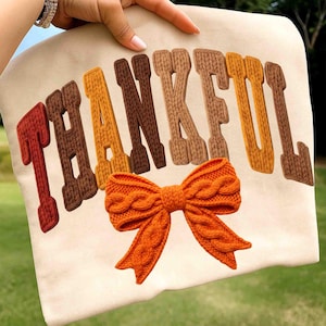 Puede incluir: Una sudadera beige con la palabra "THANKFUL" en letras texturizadas multicolores. Un lazo naranja tejido está centrado debajo del texto. Las letras son en tonos marrones y naranjas, con una textura tejida.