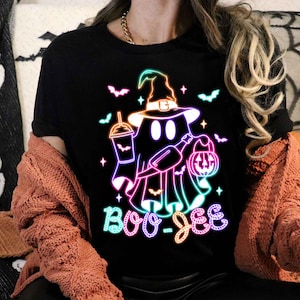 May include: A black t-shirt featuring a neon green, pink, and blue graphic of a ghost wearing a witch hat and holding a pumpkin and a drink. The text "Boo-Jee" is written below the ghost.