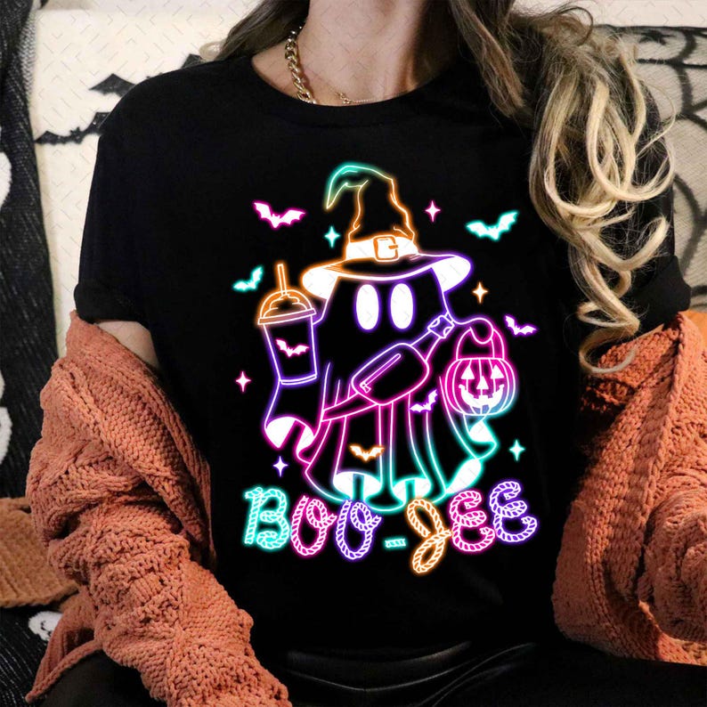 May include: A black t-shirt featuring a neon green, pink, and blue graphic of a ghost wearing a witch hat and holding a pumpkin and a drink. The text "Boo-Jee" is written below the ghost.