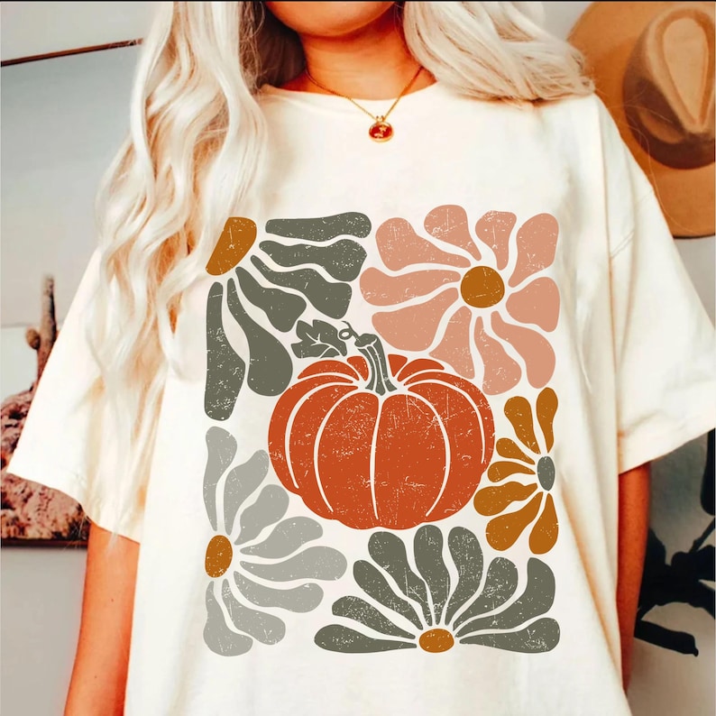 May include: Cream-colored t-shirt featuring a retro-style design with a large orange pumpkin and stylized flowers in shades of gray, brown, and pink. The shirt has short sleeves and a relaxed fit, perfect for fall.