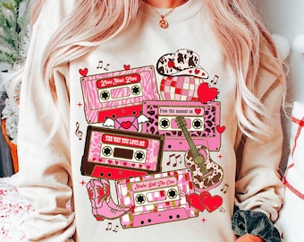 May include: A white sweatshirt with a pink and brown graphic design featuring cassette tapes with song titles "Love Your Love", "The Way You Love Me", "From This Moment On", and "You're Still The One".