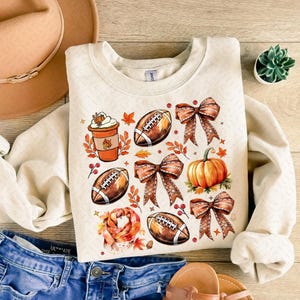 May include: A cream-colored sweatshirt with a fall-themed design featuring footballs, pumpkins, coffee cups, and bows. The design is in shades of brown, orange, and red.