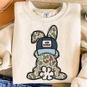 May include: A beige sweatshirt featuring a cartoon bunny wearing camouflage and a blue baseball cap with the words "egg hunter".