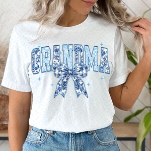 May include: White t-shirt with the word "Grandma" in blue and white floral letters with a blue and white floral bow underneath.