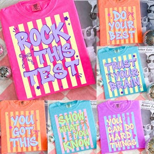 Può includere: Una collezione di magliette colorate con frasi motivazionali. Le magliette sono rosa, arancioni, blu e viola, ognuna con un messaggio unico in un font giocoso. Strisce verticali e stelle completano il design.