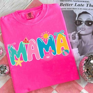 May include: Bright pink t-shirt with the word "MAMA" in colorful letters. The "M" is turquoise, the "A"s are pink and yellow, and the design includes a watermelon slice, sun, flamingo float, and beach ball.