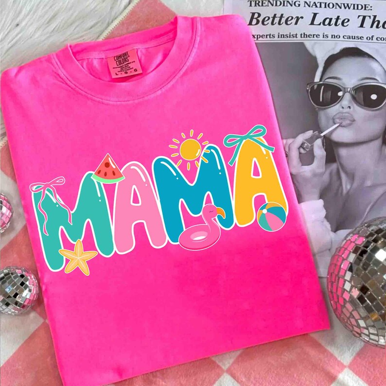 May include: Bright pink t-shirt with the word "MAMA" in colorful letters. The "M" is turquoise, the "A"s are pink and yellow, and the design includes a watermelon slice, sun, flamingo float, and beach ball.