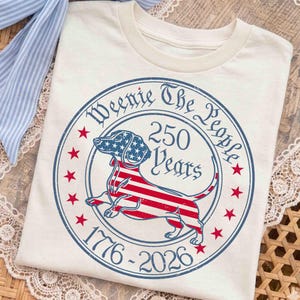 May include: A cream-colored t-shirt featuring a circular graphic with a dachshund silhouette filled with a red, white, and blue American flag pattern. The text reads Weenie The People, 250 Years, and 1776-2026, surrounded by red stars.