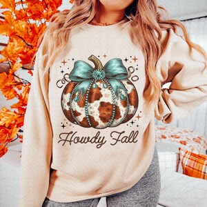 Puede incluir: Una sudadera beige con una calabaza de estampado de vaca marr&oacute;n y blanco con un lazo azul y pedrer&iacute;a. El texto "Howdy Fall" est&aacute; impreso debajo de la calabaza.