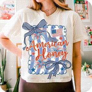 May include: Cream-coloured t-shirt with a patriotic design. The graphic features the text "American Honey" in red script, a blue and white striped background, fireworks, stars, and a small American flag. Two blue bows are also included.