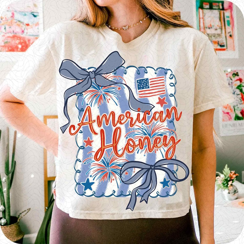 May include: Cream-coloured t-shirt with a patriotic design. The graphic features the text "American Honey" in red script, a blue and white striped background, fireworks, stars, and a small American flag. Two blue bows are also included.