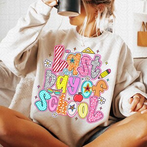 Pode incluir: Uma camisa de moletom bege claro com o texto "&Uacute;ltimo dia de aula" em letras coloridas e divertidas. As letras s&atilde;o decoradas com v&aacute;rios gr&aacute;ficos tem&aacute;ticos escolares, incluindo uma r&eacute;gua, tesoura, um l&aacute;pis, &oacute;culos de sol, uma estrela e uma ma&ccedil;&atilde;.