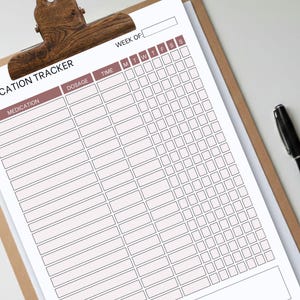 Könnte beinhalten: Ein Medikamenten-Tracker-Blatt auf einem Klemmbrett mit einer Holzklammer. Das Blatt hat eine rosa Überschrift und ein Raster zur Verfolgung der Medikamentendosierung und -zeit. Ein schwarzer Stift liegt auf der weißen Oberfläche neben dem Klemmbrett.