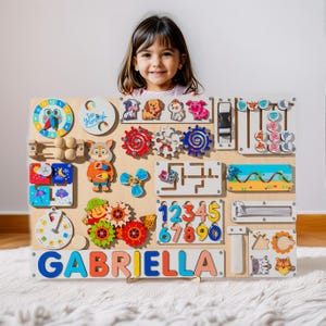 May include: A wooden activity board with various interactive elements, including a clock, gears, a maze, a zipper, a slide, and a number puzzle. The board is personalized with the name "GABRIELLA" in colorful letters. A young girl is holding the board in front of her.