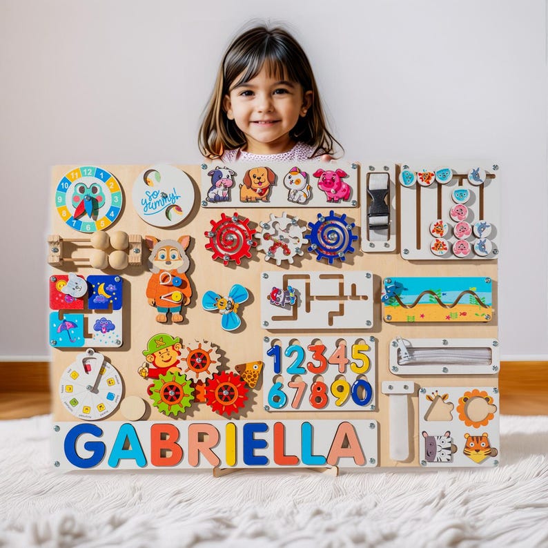 May include: A wooden activity board with various interactive elements, including a clock, gears, a maze, a zipper, a slide, and a number puzzle. The board is personalized with the name "GABRIELLA" in colorful letters. A young girl is holding the board in front of her.