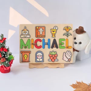 May include: A wooden puzzle board with the name "MICHAEL" spelled out in colorful letters. The board features various religious icons, including a church, a cross, a chalice, a bible, a nun, a flower, and a hand with a cross.
