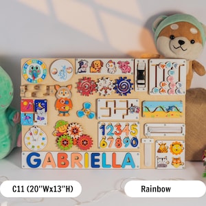 May include: A colourful wooden activity board for toddlers with various activities, including a clock, gears, a maze, numbers, a zipper, and a sliding door. The board is personalised with the name "GABRIELLA" in colourful letters. The board is 20 inches wide and 13 inches high.