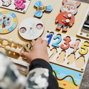 May include: Wooden activity board with colorful animal and number cutouts. Features a clock, abacus, and a spinning butterfly. The board includes the text "So Yummy!" and is designed for early childhood learning and play.