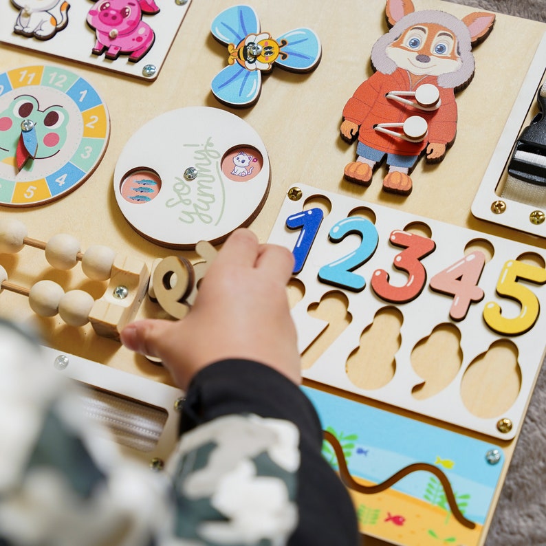May include: Wooden activity board with colorful animal and number cutouts. Features a clock, abacus, and a spinning butterfly. The board includes the text "So Yummy!" and is designed for early childhood learning and play.