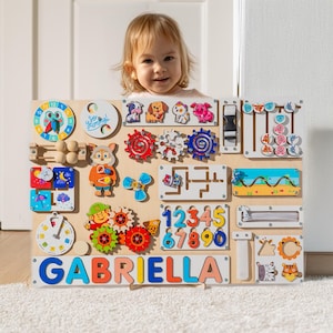 May include: A wooden activity board with various interactive elements for children, including gears, a clock, a maze, a zipper, a slider, and a shape sorter. The board is painted in bright colours and features a nameplate with the name "GABRIELLA".