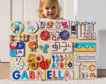 Puede incluir: Un tablero de actividades de madera con varios elementos interactivos para niños, incluyendo engranajes, un reloj, un laberinto, una cremallera, un deslizador y un clasificador de formas. El tablero está pintado en colores brillantes y presenta una placa de nombre con el nombre "GABRIELLA".