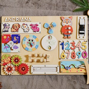 May include: Wooden activity board featuring the name "ANDRIANA" and various interactive elements. Includes animal figures, a clock, numbers, gears, a zipper, and a buckle. The board is designed for early childhood learning and play.