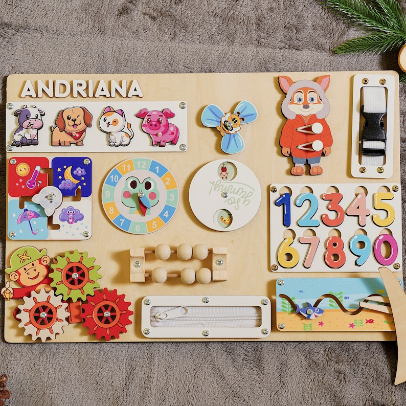 May include: Wooden activity board featuring the name "ANDRIANA" and various interactive elements. Includes animal figures, a clock, numbers, gears, a zipper, and a buckle. The board is designed for early childhood learning and play.