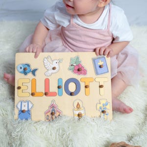 Puede incluir: Un rompecabezas de madera personalizado con el nombre "ELLIOTT" en letras de colores. El rompecabezas presenta varios s&iacute;mbolos religiosos, incluyendo una paloma, una cruz, una iglesia, un pez, una vela y una biblia.