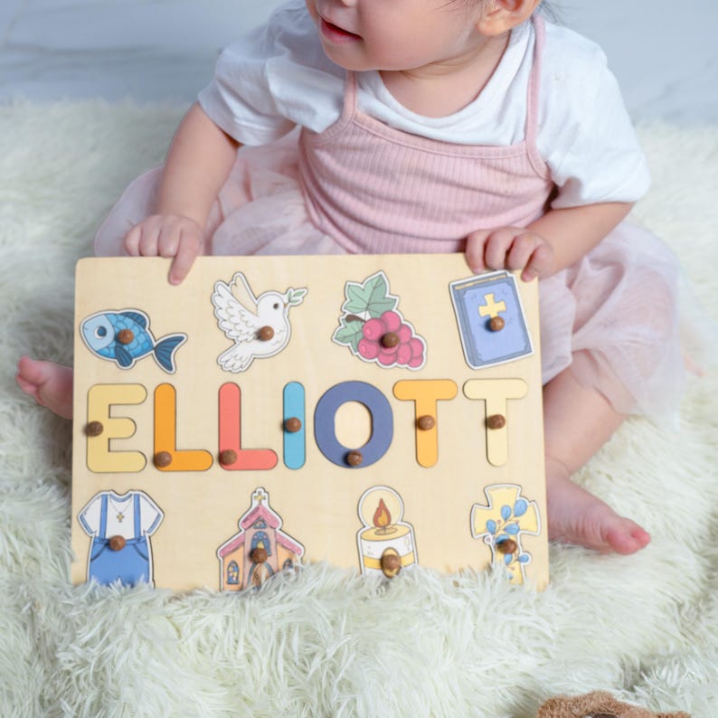 Puede incluir: Un rompecabezas de madera personalizado con el nombre "ELLIOTT" en letras de colores. El rompecabezas presenta varios s&iacute;mbolos religiosos, incluyendo una paloma, una cruz, una iglesia, un pez, una vela y una biblia.