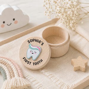 May include: A small, round, light-colored wooden box with a removable lid. The lid features a cartoon tooth graphic with a swirl of toothpaste on top and the black text "SOPHIA'S FIRST TOOTH" printed around the illustration in a circular layout.