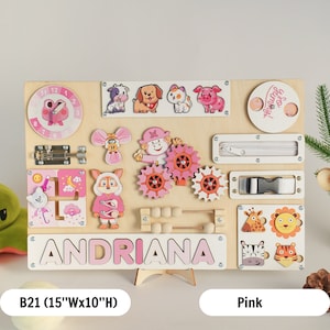 May include: A pink and white wooden busy board with a variety of activities for children, including a clock, a zipper, a buckle, a latch, a maze, a gear set, an abacus, and a name puzzle. The board is personalised with the name "ANDRIANA".