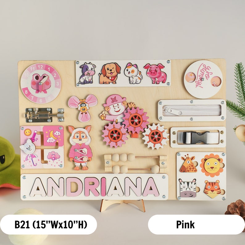 May include: A pink and white wooden busy board with a variety of activities for children, including a clock, a zipper, a buckle, a latch, a maze, a gear set, an abacus, and a name puzzle. The board is personalised with the name "ANDRIANA".