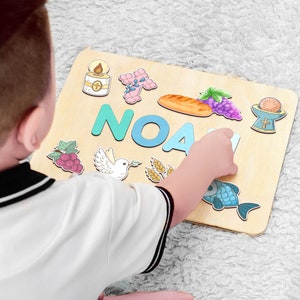 Puede incluir: Un tablero de madera con el nombre "NOAH" escrito en letras de colores. El tablero est&aacute; decorado con varios s&iacute;mbolos religiosos, incluyendo una paloma, una cruz, un c&aacute;liz, uvas, pan y un pez.