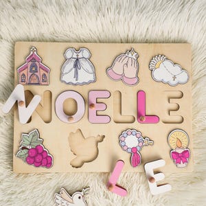 May include: A wooden puzzle with the name "NOELLE" spelled out in pink and white letters. The puzzle includes various religious symbols, such as a church, a dove, a rosary, and a candle.