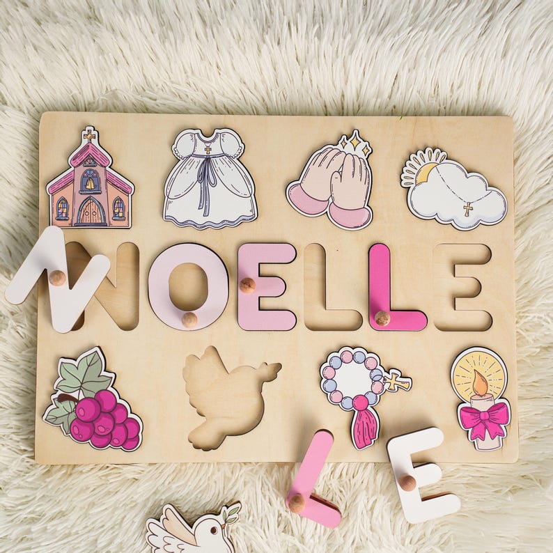 May include: A wooden puzzle with the name "NOELLE" spelled out in pink and white letters. The puzzle includes various religious symbols, such as a church, a dove, a rosary, and a candle.