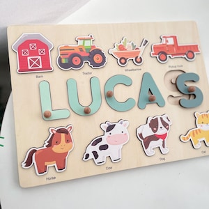 May include: A wooden puzzle board with a light brown background. The puzzle features the name "LUCAS" in large, light blue letters. There are also images of a barn, tractor, wheelbarrow, pickup truck, horse, cow, dog, and cat. Each image has a corresponding label underneath.