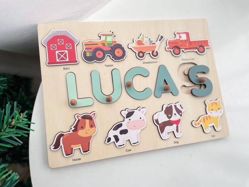 May include: A wooden puzzle board with a light brown background. The puzzle features the name "LUCAS" in large, light blue letters. There are also images of a barn, tractor, wheelbarrow, pickup truck, horse, cow, dog, and cat. Each image has a corresponding label underneath.