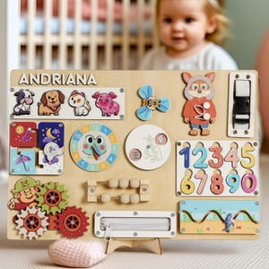 May include: Wooden activity board with various interactive elements for toddlers. Features animal figures, a clock, gears, numbers, a zipper, and the name "ANDRIANA". Includes a small abacus and a buckle. The board is designed for educational play.